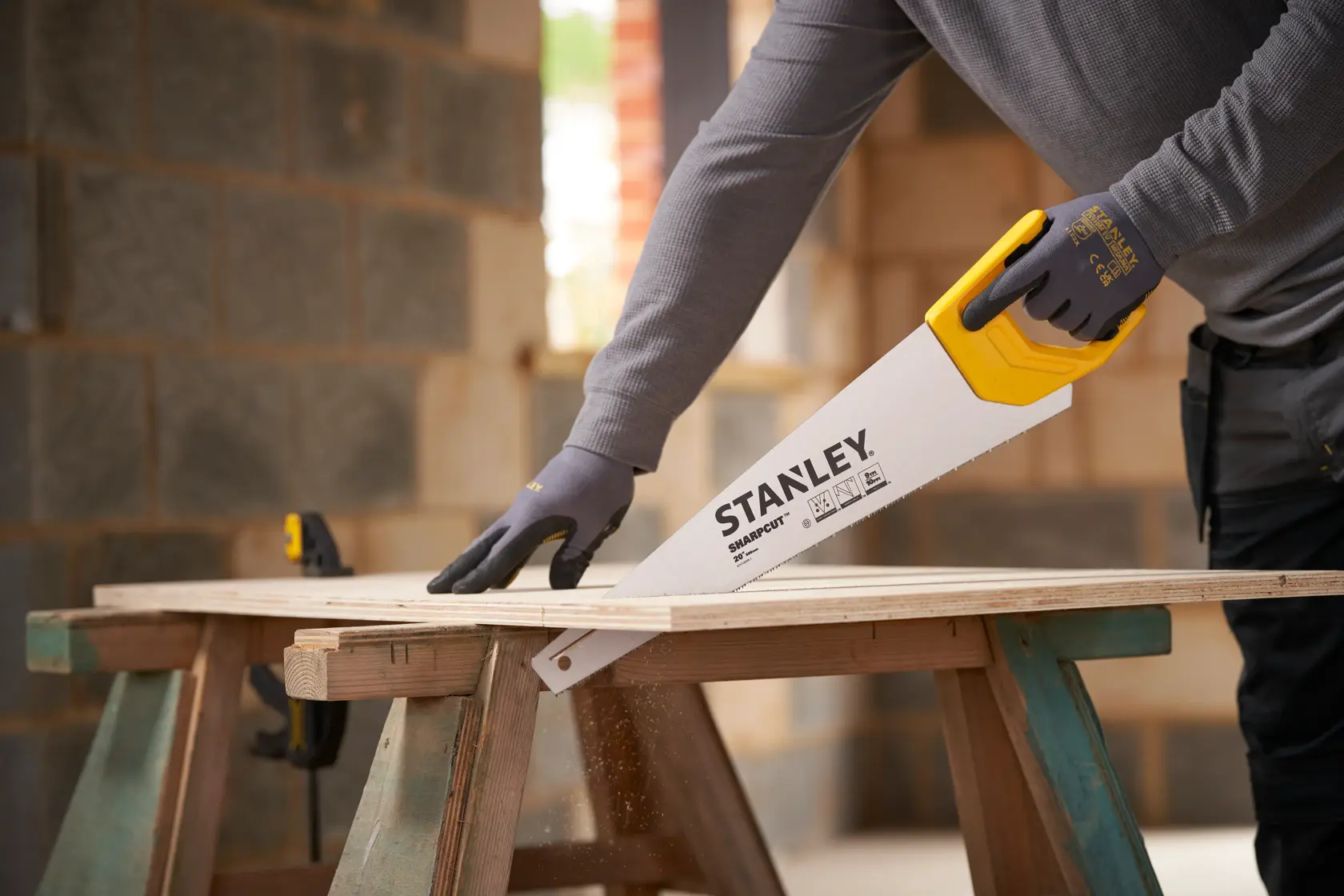 Stanley STHT20375-1 hand saw with yellow handle cutting wood plank on sawhorse, worker wearing gloves.