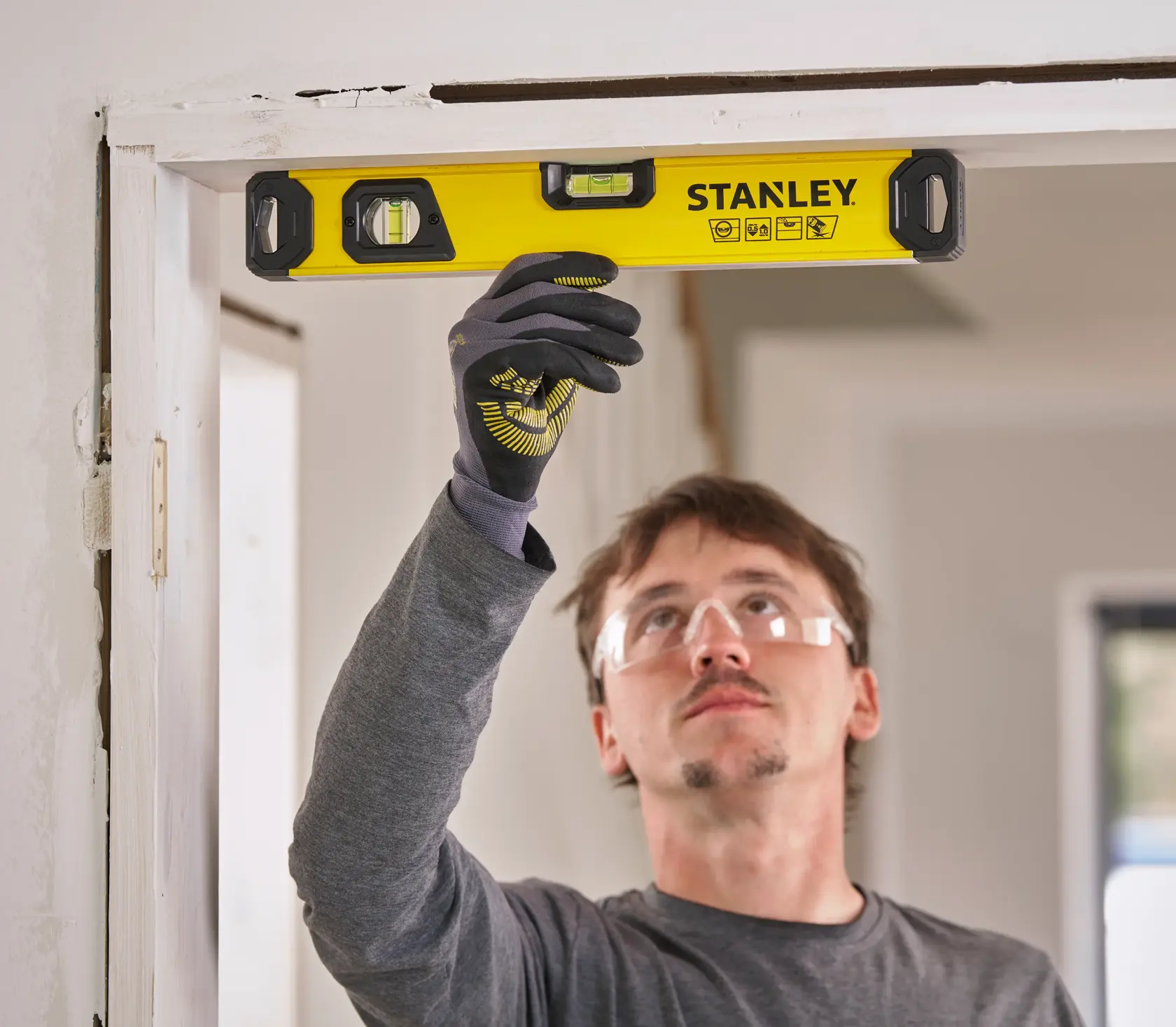 A person wearing a grey shirt and protective gloves is using a yellow STANLEY spirit level to check the alignment of a door frame in a home interior.
