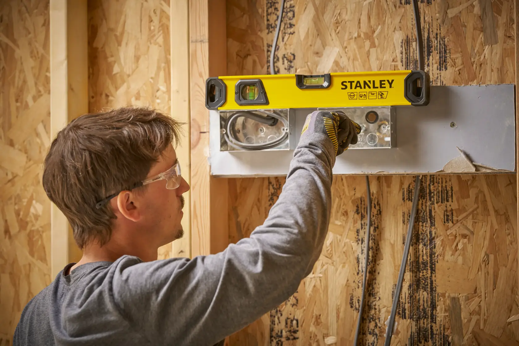 A person uses a STANLEY spirit level (model STHT41016-0) to check the alignment of a metal fixture on a wooden wall under construction.