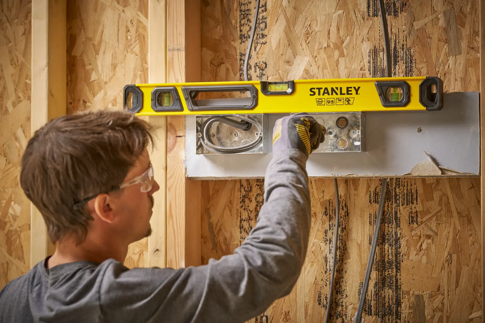 A person using a yellow STANLEY spirit level on a wooden wall frame during construction. The person's face is blurred for privacy. The spirit level is being used to align an electrical fixture.