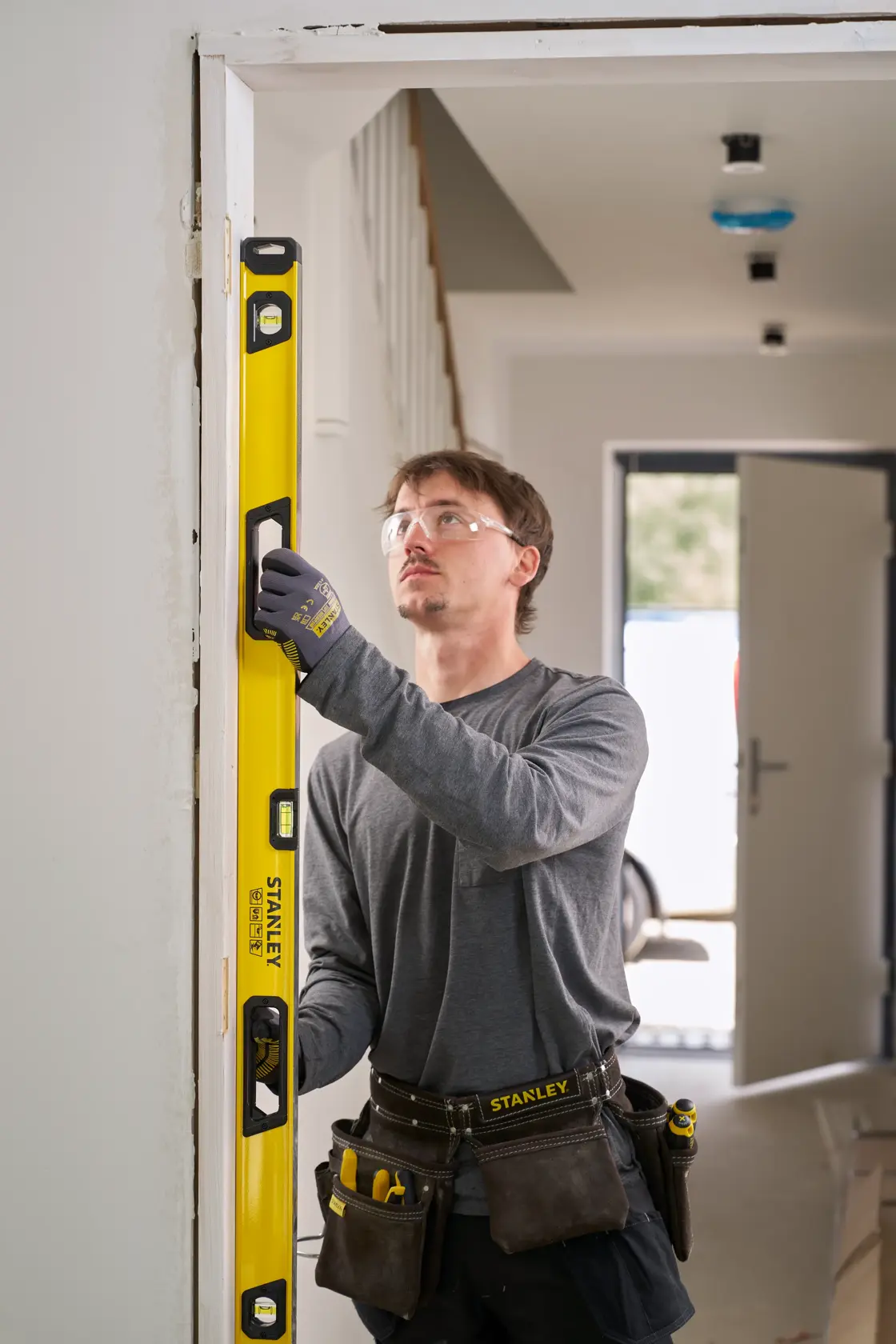 Person using a yellow STANLEY spirit level to measure the vertical alignment of a door frame. The individual is wearing work gloves, a tool belt, and casual work attire in an indoor setting.