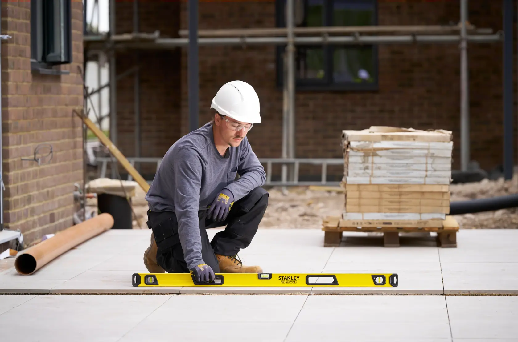 A person wearing a white hard hat and gloves is using a yellow Stanley spirit level to check the alignment of floor tiles at a construction site.