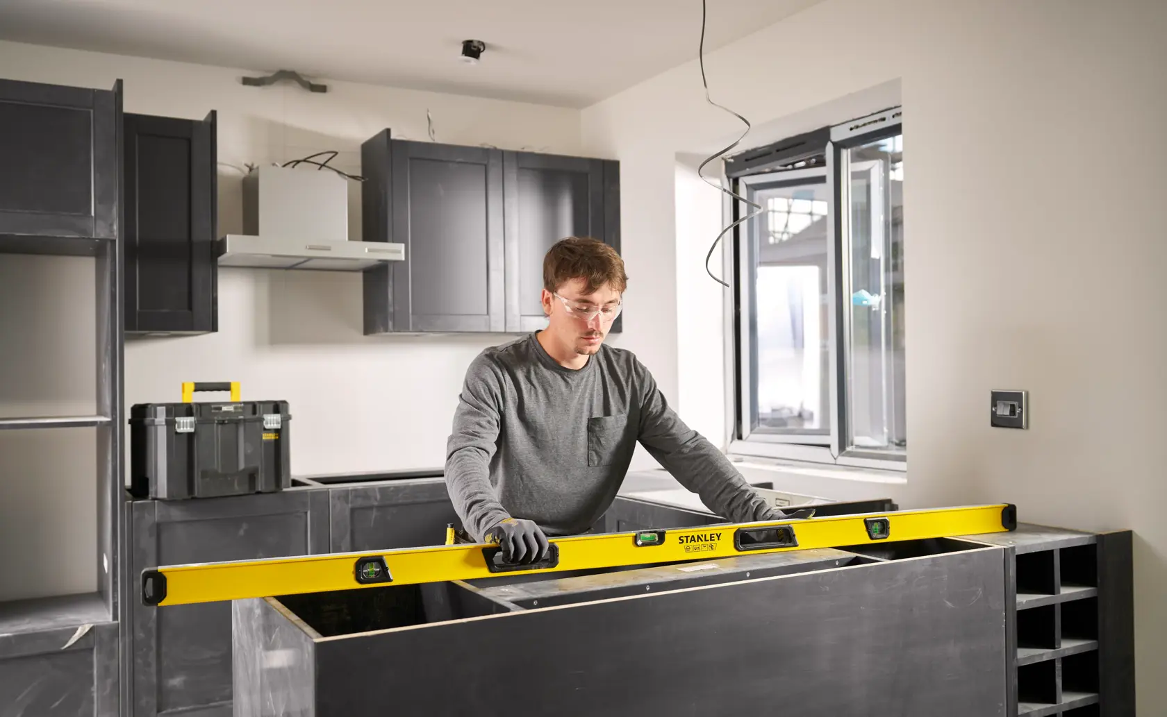 A person is using a yellow STANLEY spirit level to check the alignment of a countertop in a modern kitchen under construction. Black cabinets and a toolbox are visible in the background. The person's face is blurred.