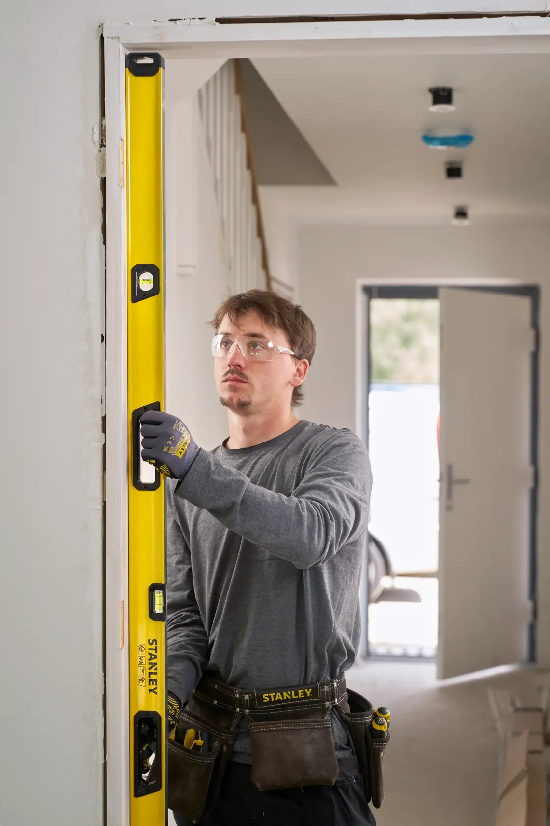 A person wearing work gloves and a tool belt is using a yellow STANLEY spirit level to check the vertical alignment of a door frame in a house under construction or renovation.