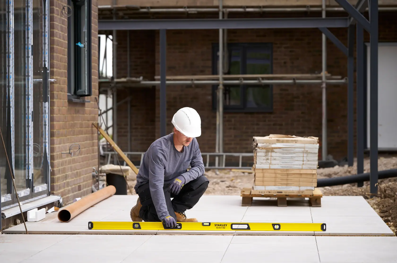 A construction worker wearing a white helmet and gloves is using a yellow STANLEY spirit level to check the alignment of outdoor pavement tiles on a building site.