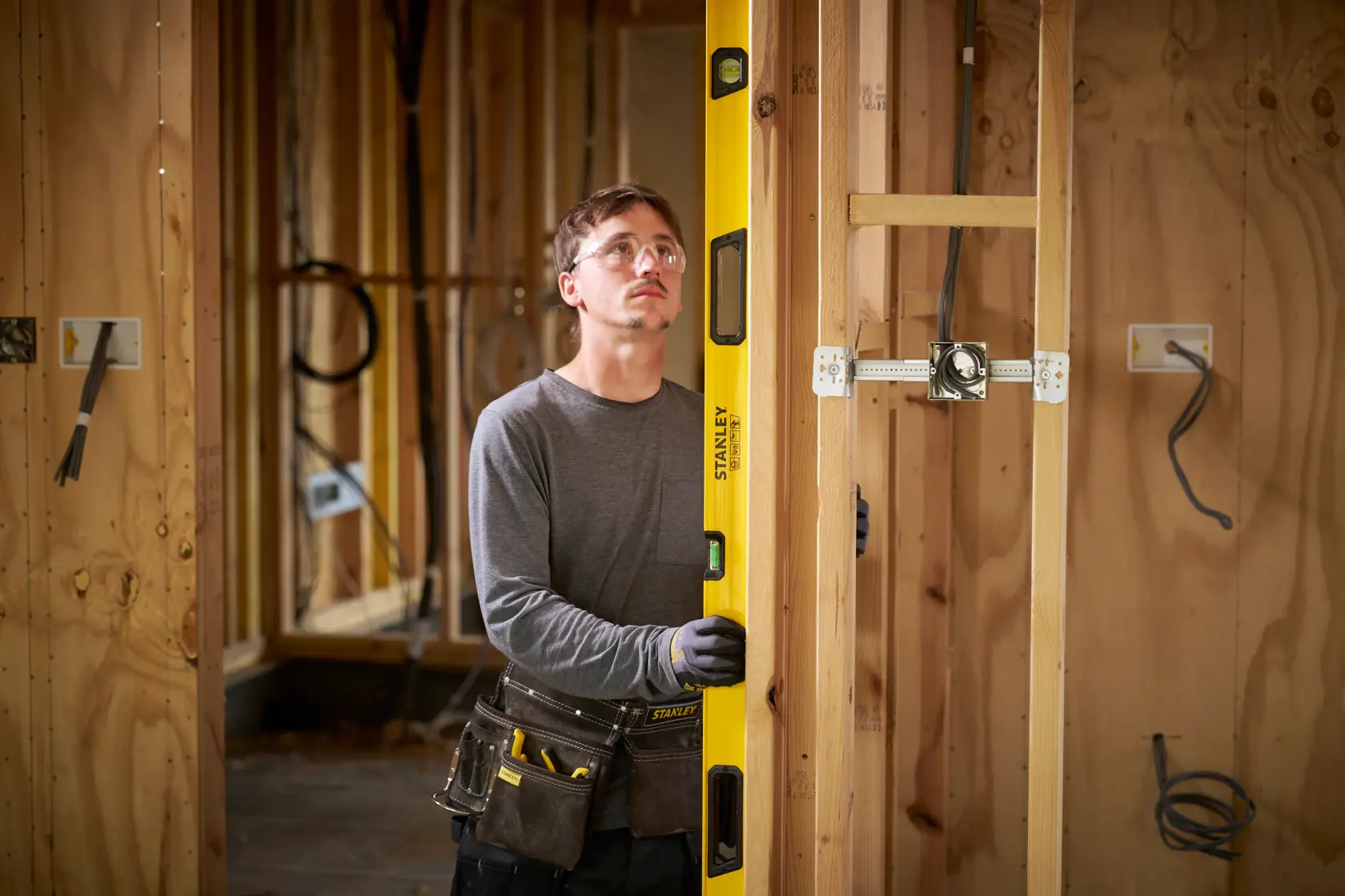 A person wearing work gloves and a tool belt is using a yellow Stanley spirit level to measure the vertical alignment of a wooden frame in a construction setting. The background shows unfinished wooden walls and electrical wiring.