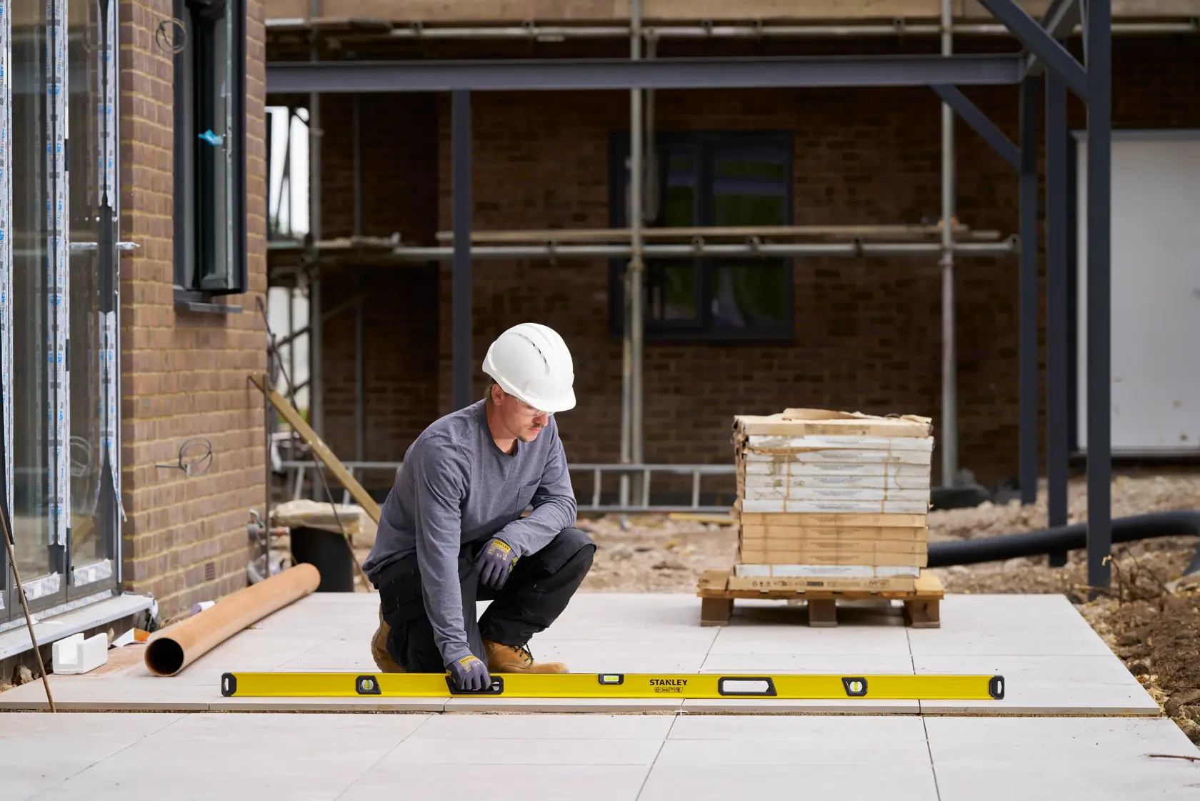A construction worker wearing a white hard hat and gloves is using a long yellow STANLEY spirit level (model STHT41078-0) to check the alignment of outdoor tiled flooring on a construction site. Building materials and scaffolding are visible in the background.