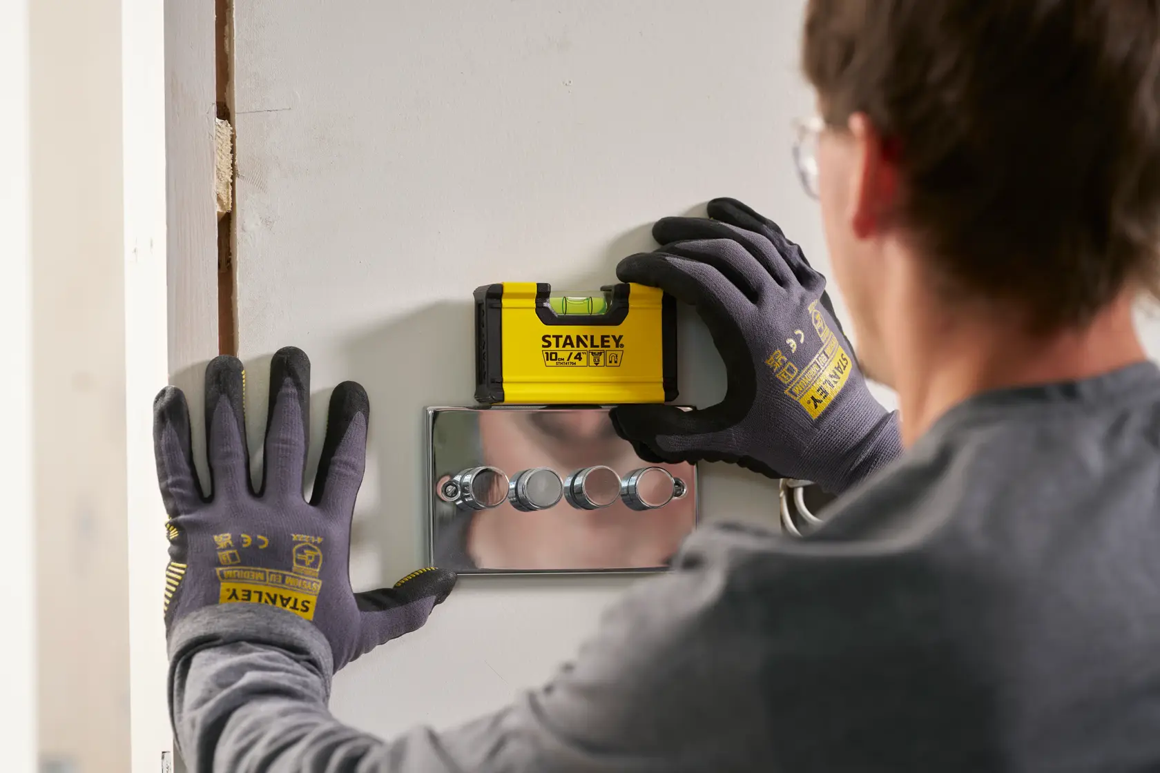 A person wearing Stanley branded gloves uses a yellow STANLEY spirit level tool (model STHT41704-0) to check alignment above a set of wall switches.