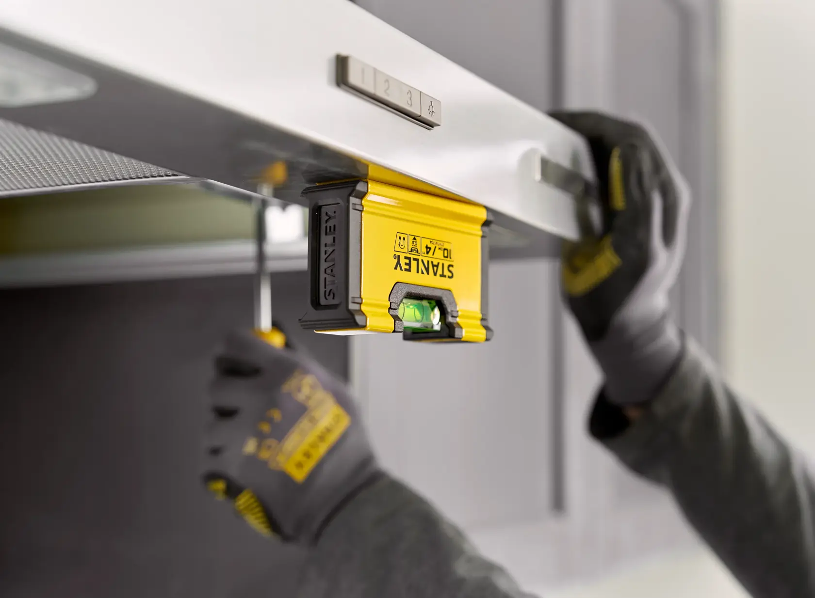 A person wearing gloves uses a yellow STANLEY spirit level (model STHT41704-0) to align a metallic surface, likely during a home improvement or installation task.
