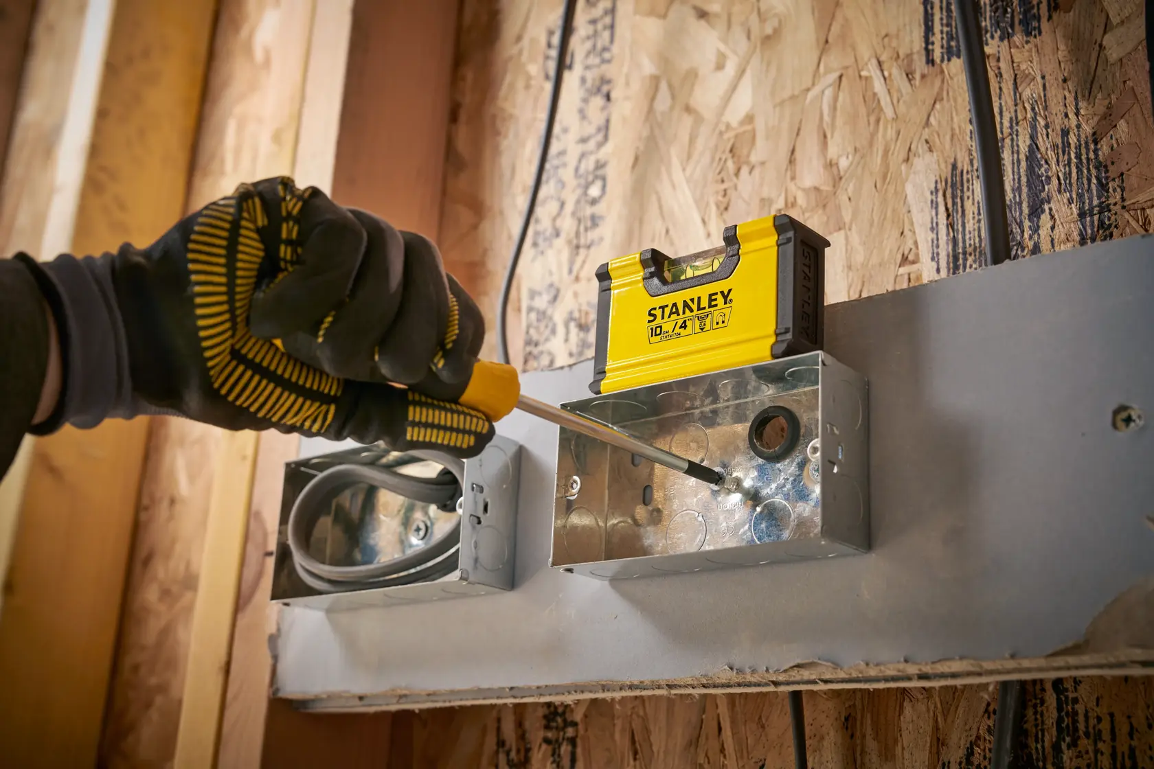A gloved hand using a screwdriver to install or adjust a metal electrical box. A yellow STANLEY tool container is positioned on top of the box. The background shows a wooden wall under construction.
