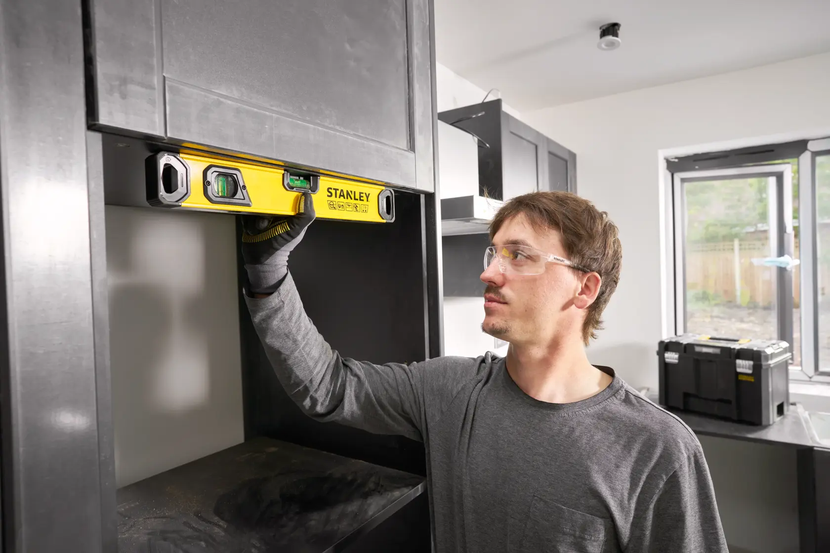 A person using a STANLEY spirit level (model STHT41716-0) to check the alignment of a shelf inside a modern kitchen or workshop. The person's face is blurred for privacy. There are black cabinets and a tool box visible in the background.