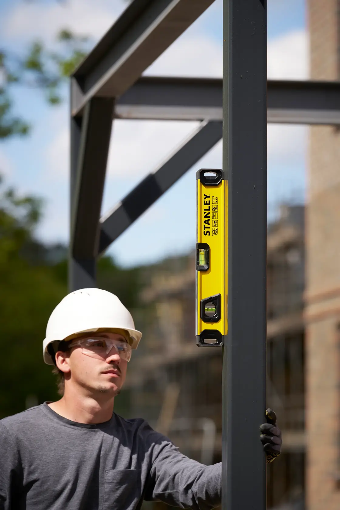 A person wearing a white safety helmet and grey shirt uses a yellow STANLEY spirit level (model STHT41716-0) to check the vertical alignment of a metal beam at an outdoor construction site. The person's face is blurred.