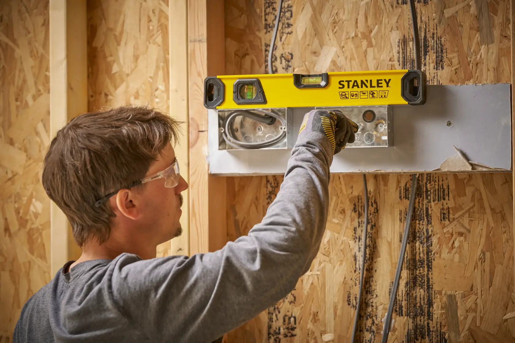 A person using a STANLEY spirit level to check the alignment of an electrical box on a wooden wall during construction. The person is wearing a grey shirt and work gloves.