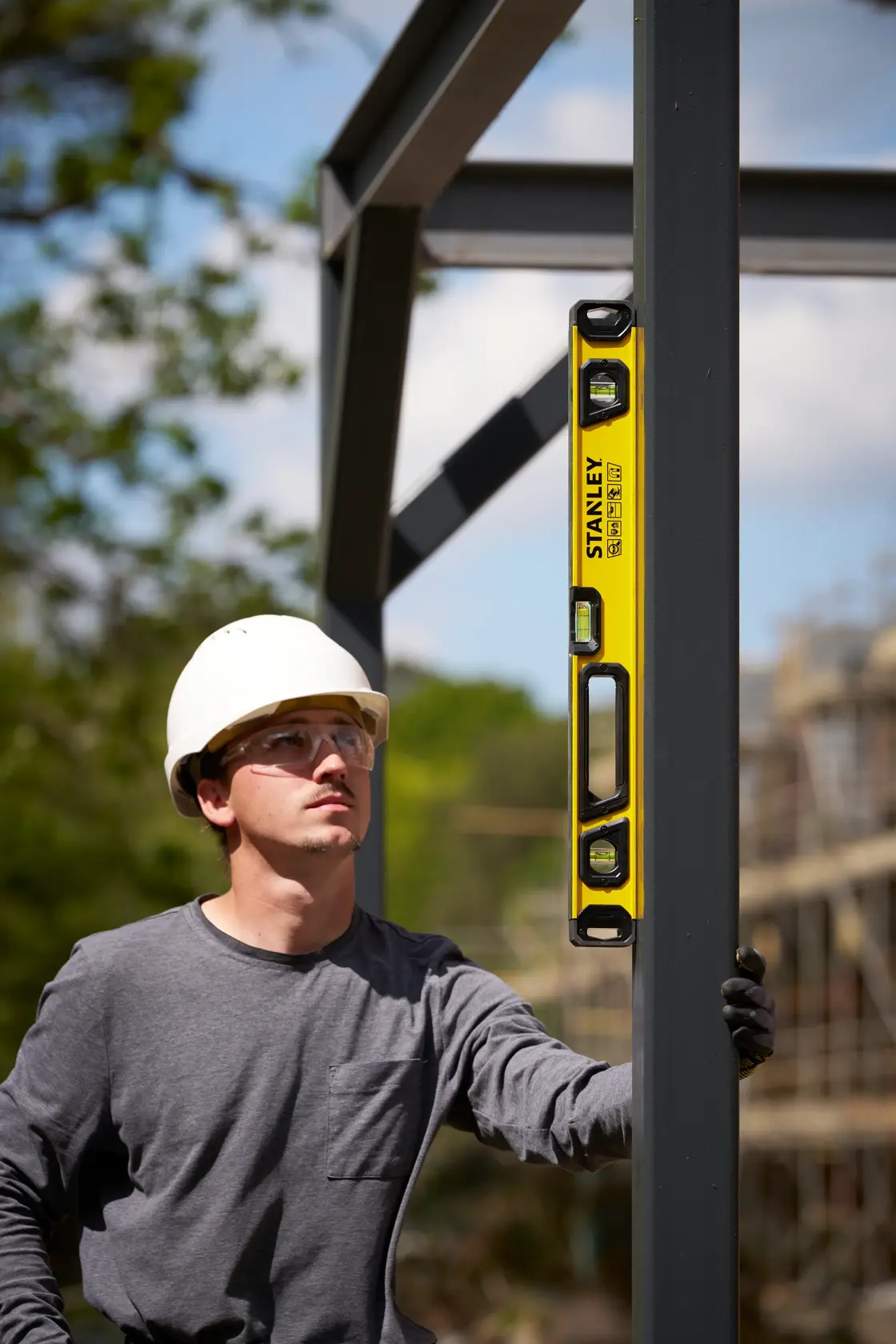 A person wearing a white safety helmet uses a yellow STANLEY spirit level to check the alignment of a vertical metal beam at a construction site. The person's face is blurred.