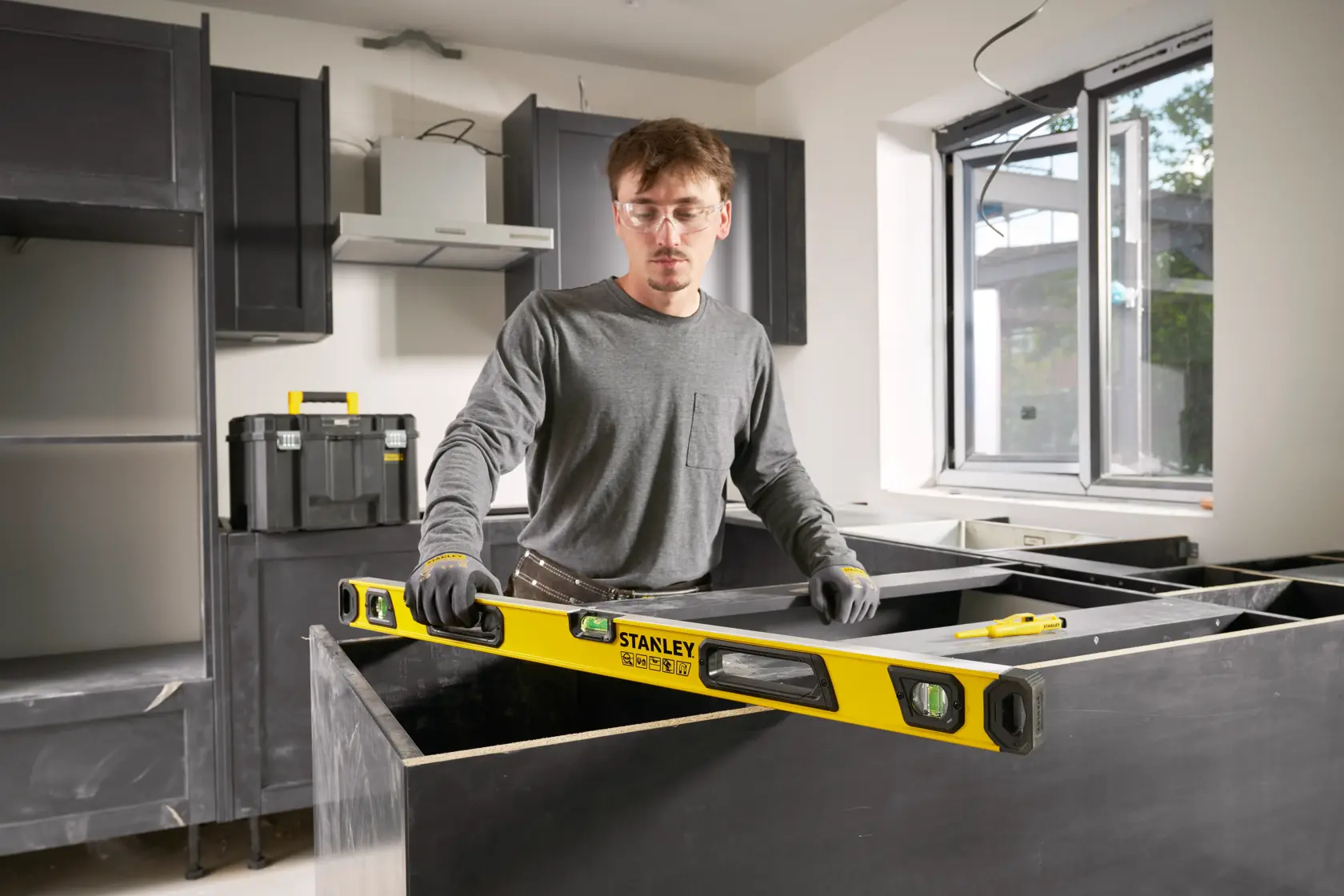 Person using a yellow STANLEY spirit level (model STHT41736-0) in a kitchen under construction. The individual is wearing gloves and grey clothing, aligning a countertop. Construction tools and materials are visible in the background.