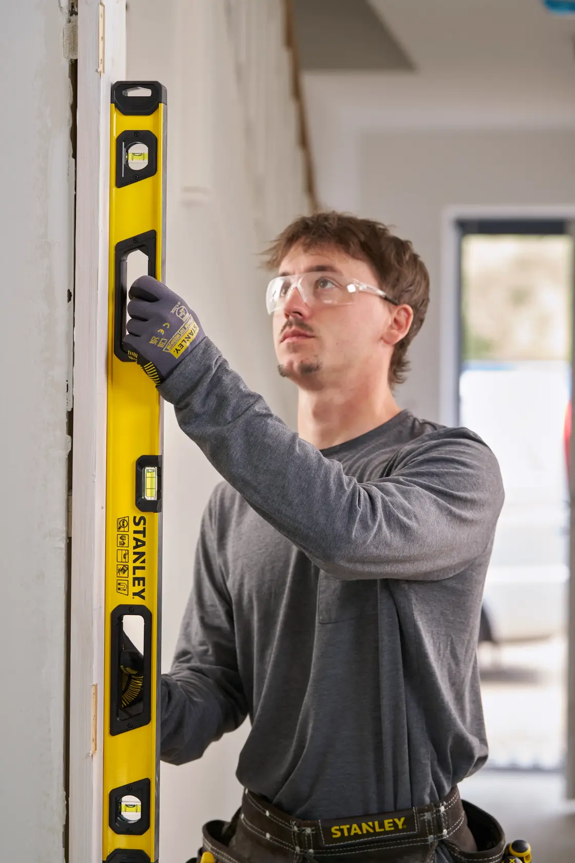 A person wearing work gloves and a grey shirt uses a yellow Stanley spirit level to check the vertical alignment of a door frame inside a building. The Stanley branding and measurement markings are visible on the tool.