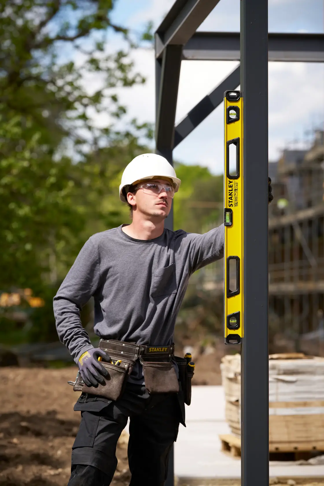 A construction worker using a yellow STANLEY spirit level (SKU: STHT41736-0) to check the alignment of a vertical metal beam at an outdoor construction site. The worker is wearing a hard hat, gray shirt, gloves, and a tool belt. The background features trees and construction materials.