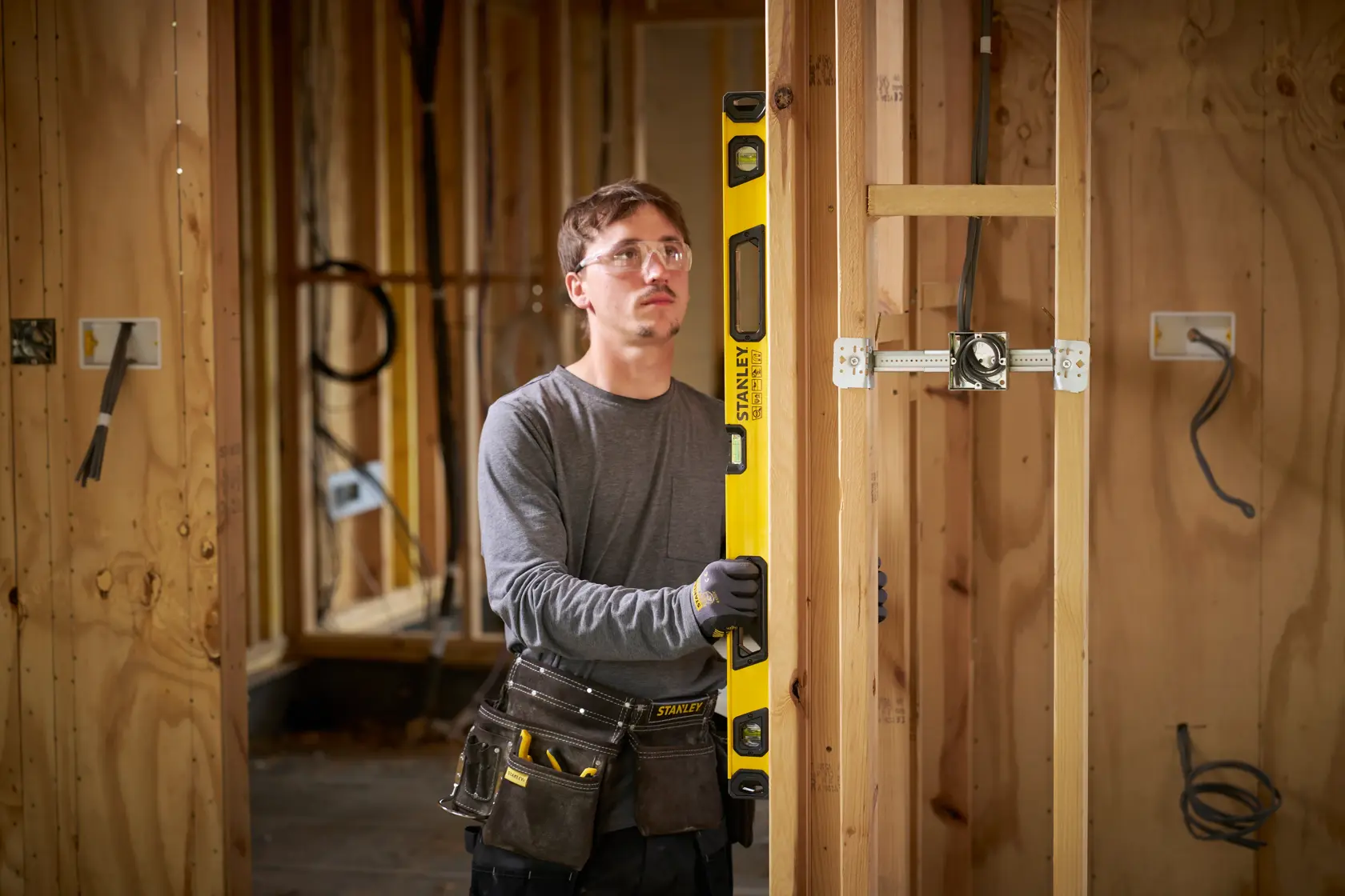A person with their face blurred is holding a yellow STANLEY spirit level against a wooden wall frame on a construction site. The person is wearing work gloves and a tool belt with various tools.
