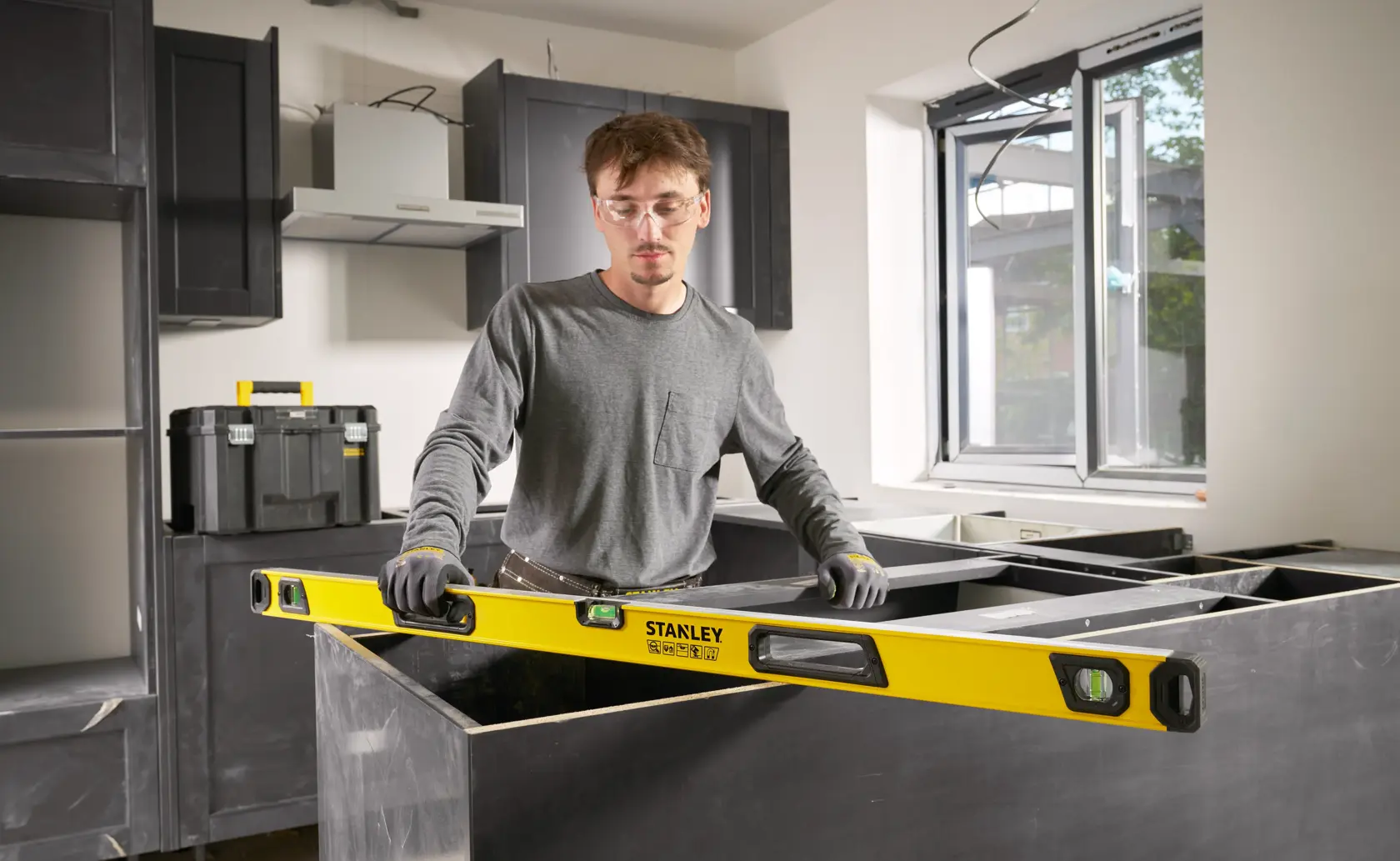 Person using a Stanley spirit level to measure and align a countertop in a kitchen under construction, with toolboxes and cabinets visible in the background.