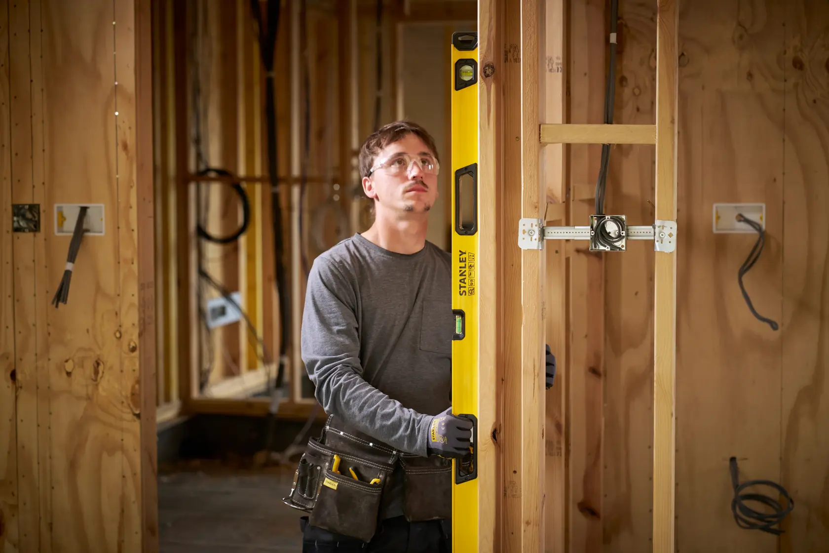A person wearing work gloves and a tool belt uses a yellow STANLEY spirit level to check the vertical alignment of a wooden stud frame in a construction site. The background shows unfinished wooden walls and electrical wiring.