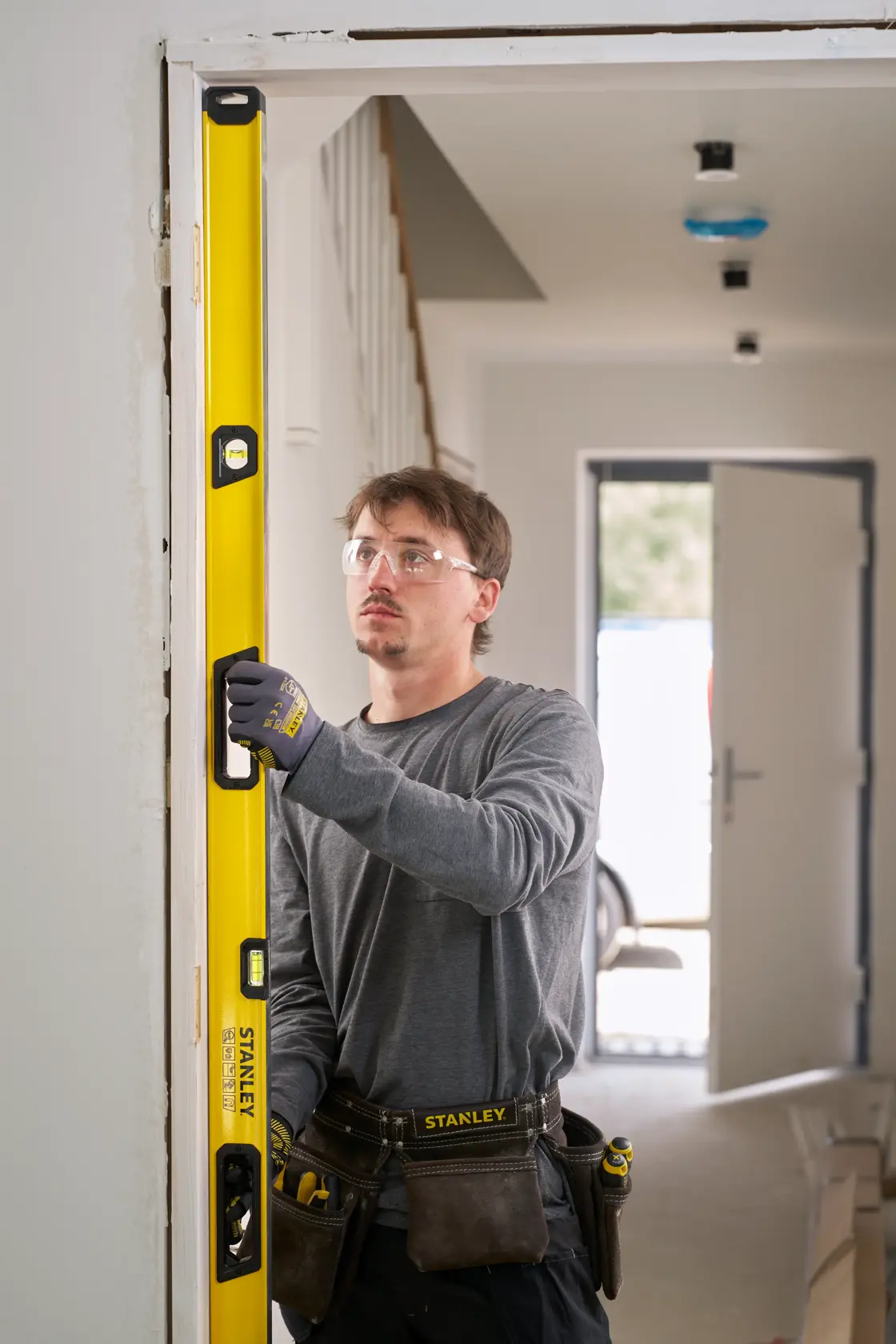 A person wearing work gloves and a tool belt is using a yellow STANLEY spirit level to check the vertical alignment of a door frame in an indoor setting.
