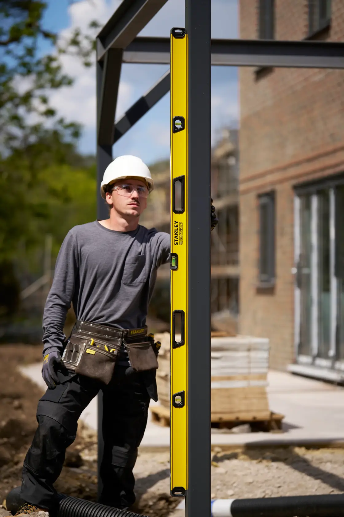 A person wearing a white safety helmet and a tool belt uses a yellow STANLEY spirit level to check the vertical alignment of a metal beam at an outdoor construction site.