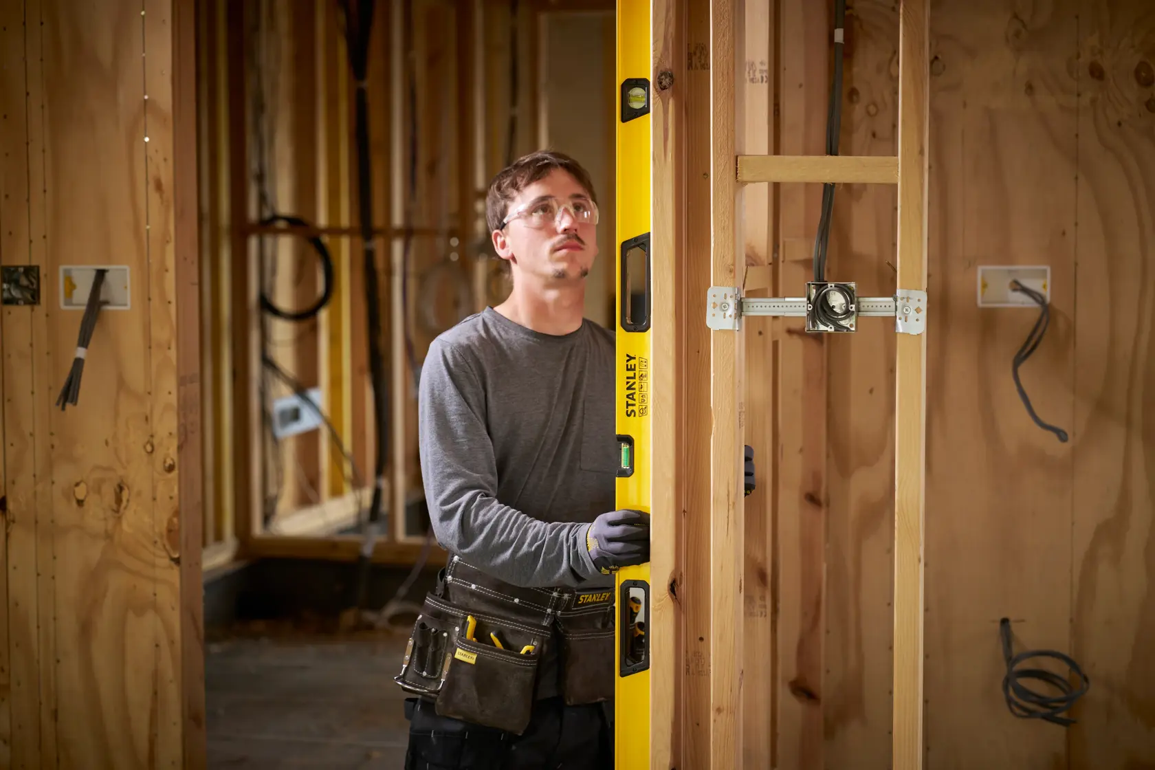 A person with a blurred face using a yellow STANLEY spirit level to check the vertical alignment of wooden framing at a construction site. The individual is wearing work gloves and a tool belt, and the surroundings show unfinished wooden walls and electrical wiring.