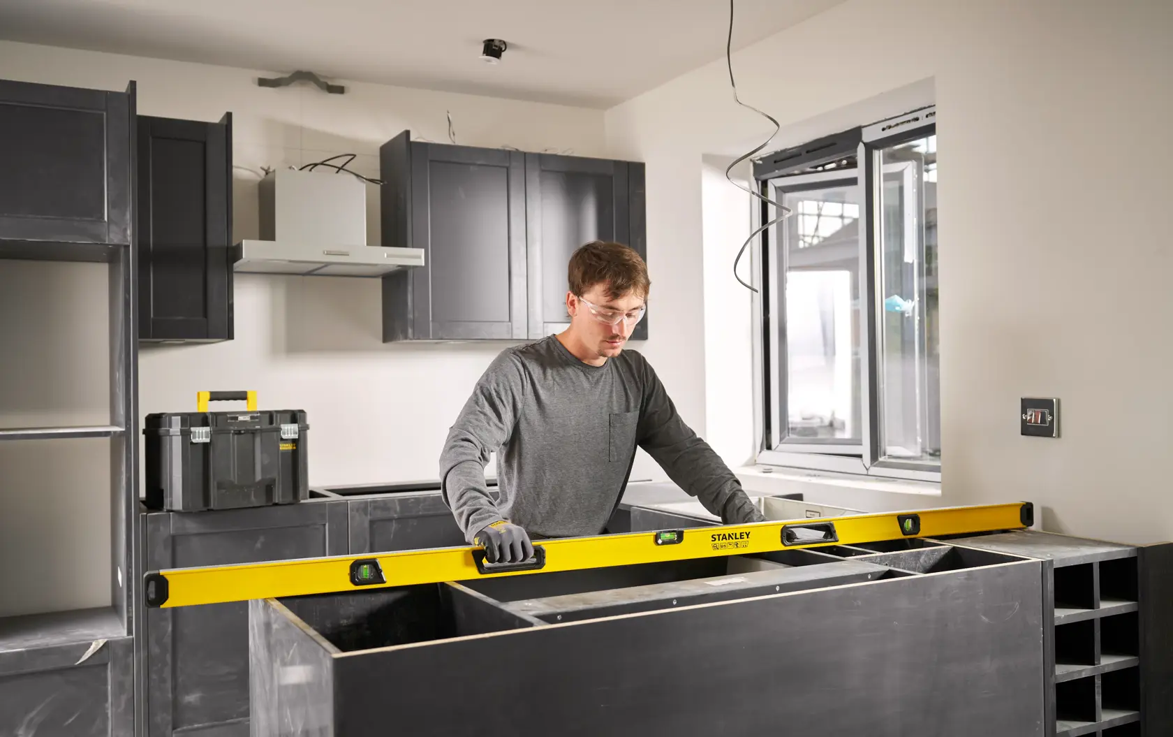 A person using a yellow STANLEY spirit level while working on a kitchen installation. The individual is wearing gloves and is measuring a countertop in a modern kitchen setting.
