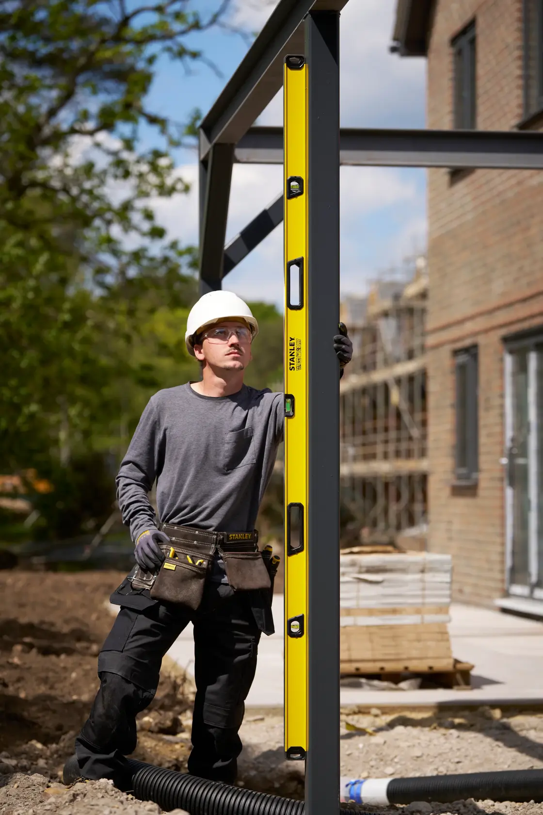 A construction worker in safety gear uses a yellow STANLEY spirit level (model STHT41778-0) to check the alignment of a vertical metal beam on an outdoor building site.