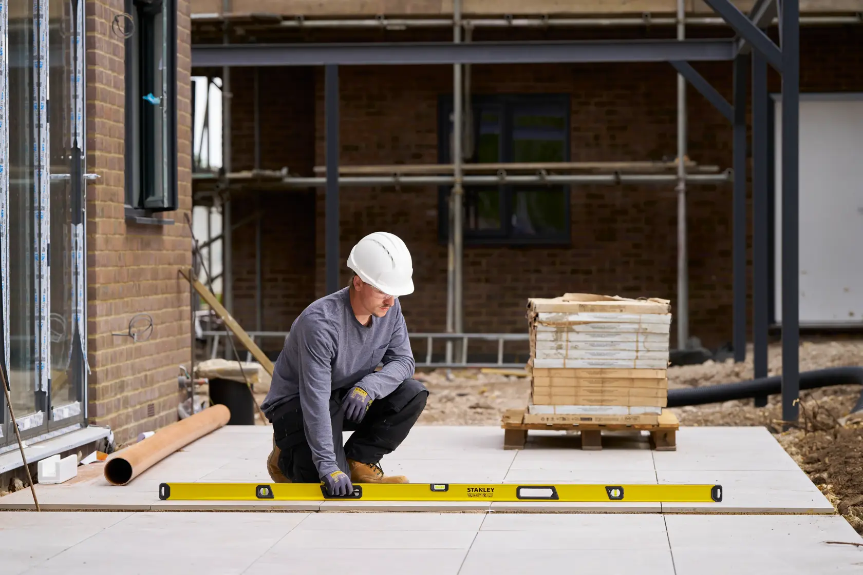 A construction worker wearing a white hard hat and gloves is kneeling outdoors, using a long yellow STANLEY spirit level to check the alignment of paving tiles. There are construction materials and scaffolding in the background.