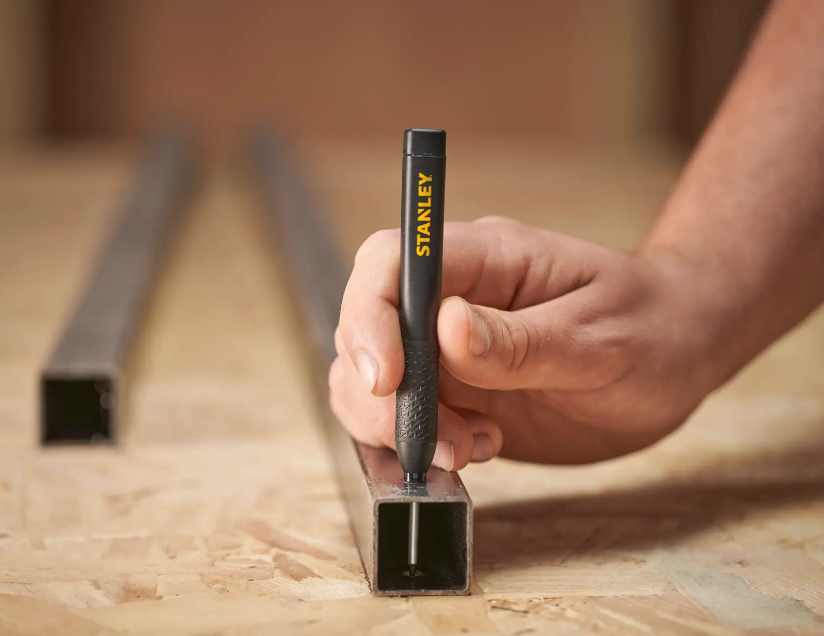 Hand using a Stanley marking pen to write on a metal tube placed on a wooden work surface.