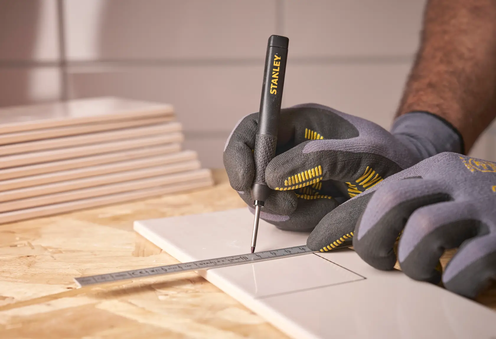 Gloved hands use a STANLEY marking tool and ruler to draw lines on a tile for precise measurement.