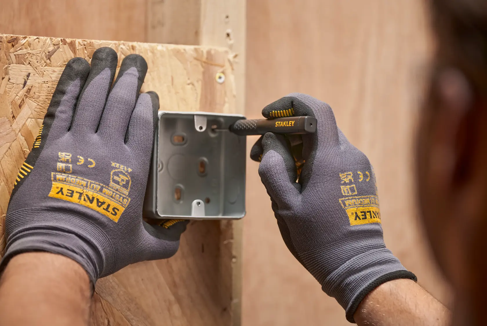 Hands with STANLEY gloves mark a metal box on wood using a STANLEY pencil during construction.