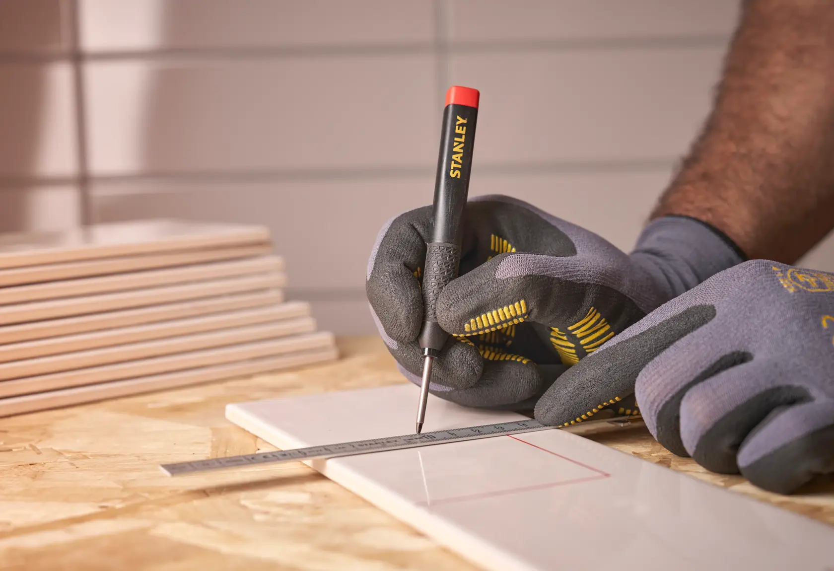 Gloved hands mark a tile with a STANLEY tool and metal ruler on a wooden surface.