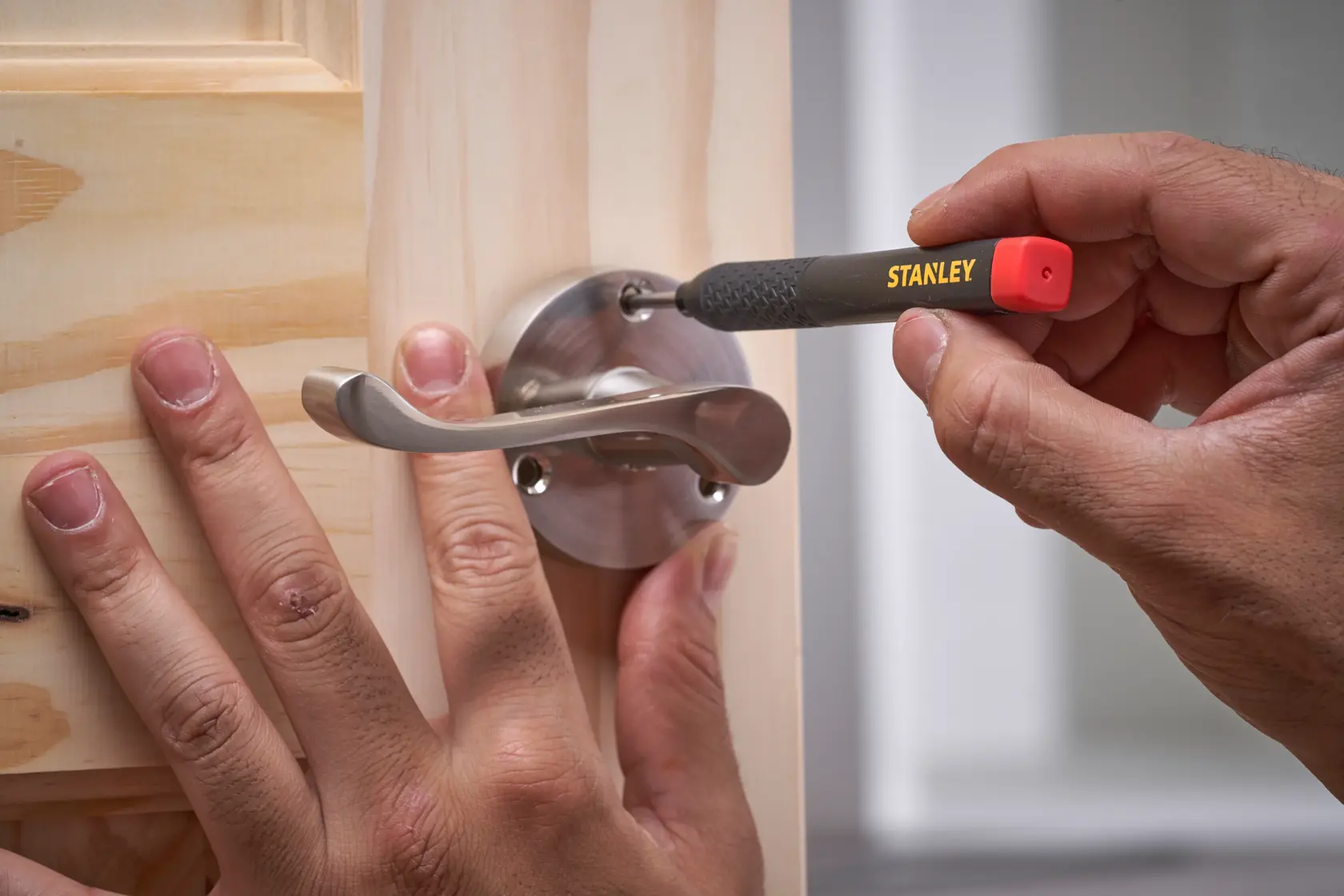 A STANLEY tool is used to tighten a screw on a metal door handle attached to a wooden door.