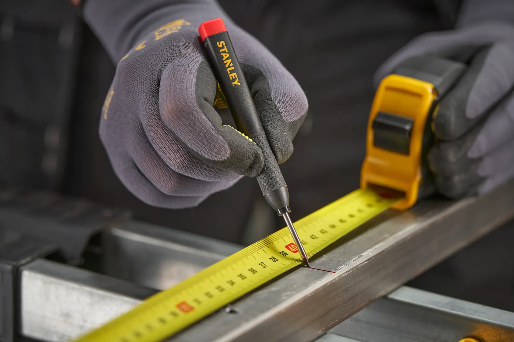 Gloved hands use a Stanley marking tool and tape measure to mark measurements on a metal surface.