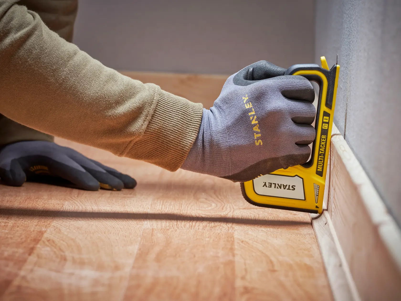Stanley STHT70440-0 manual stapler in use, yellow and black, held by gloved hand against baseboard.