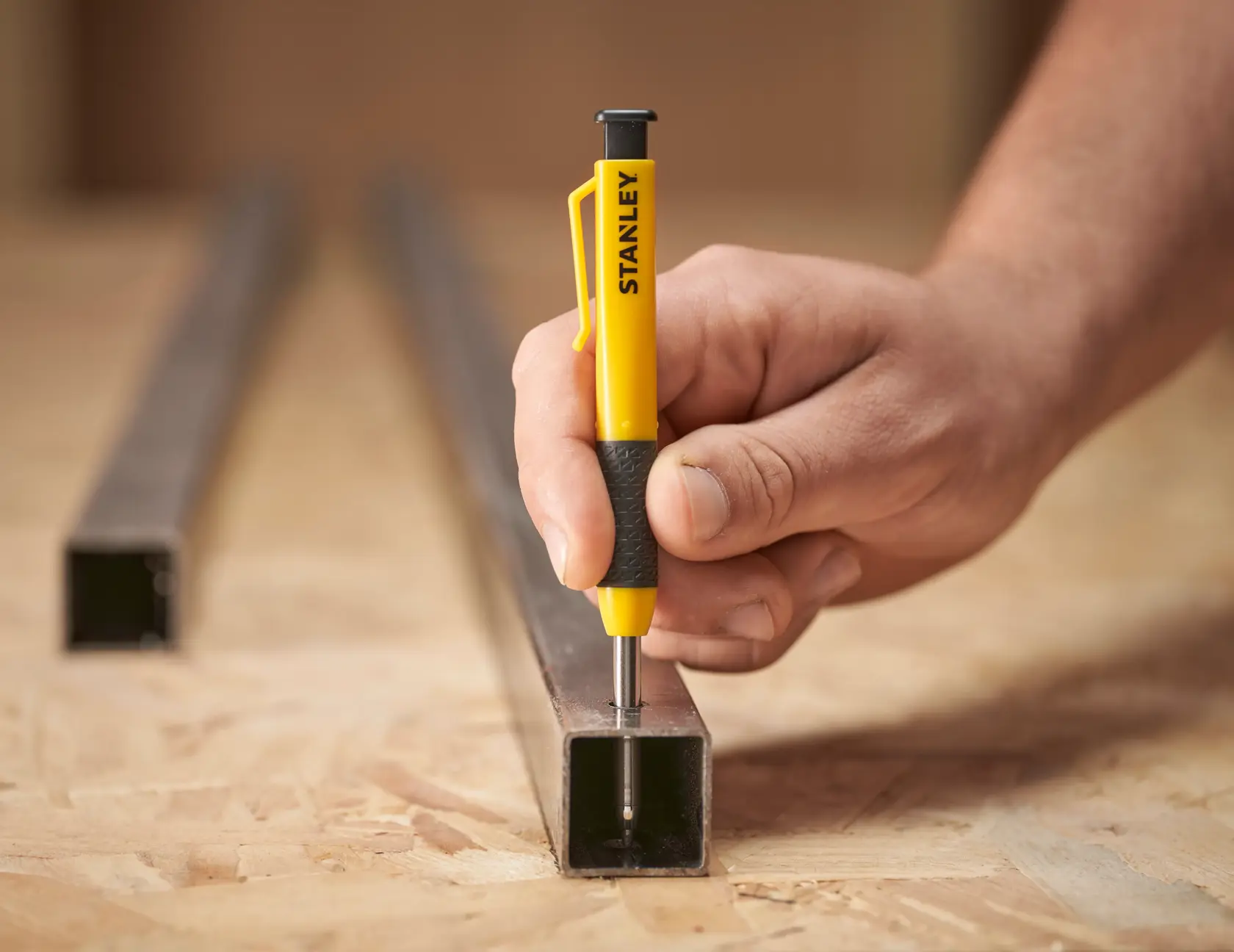 Hand using a STANLEY spring-loaded center punch on a metal square tube placed on a wooden work surface.