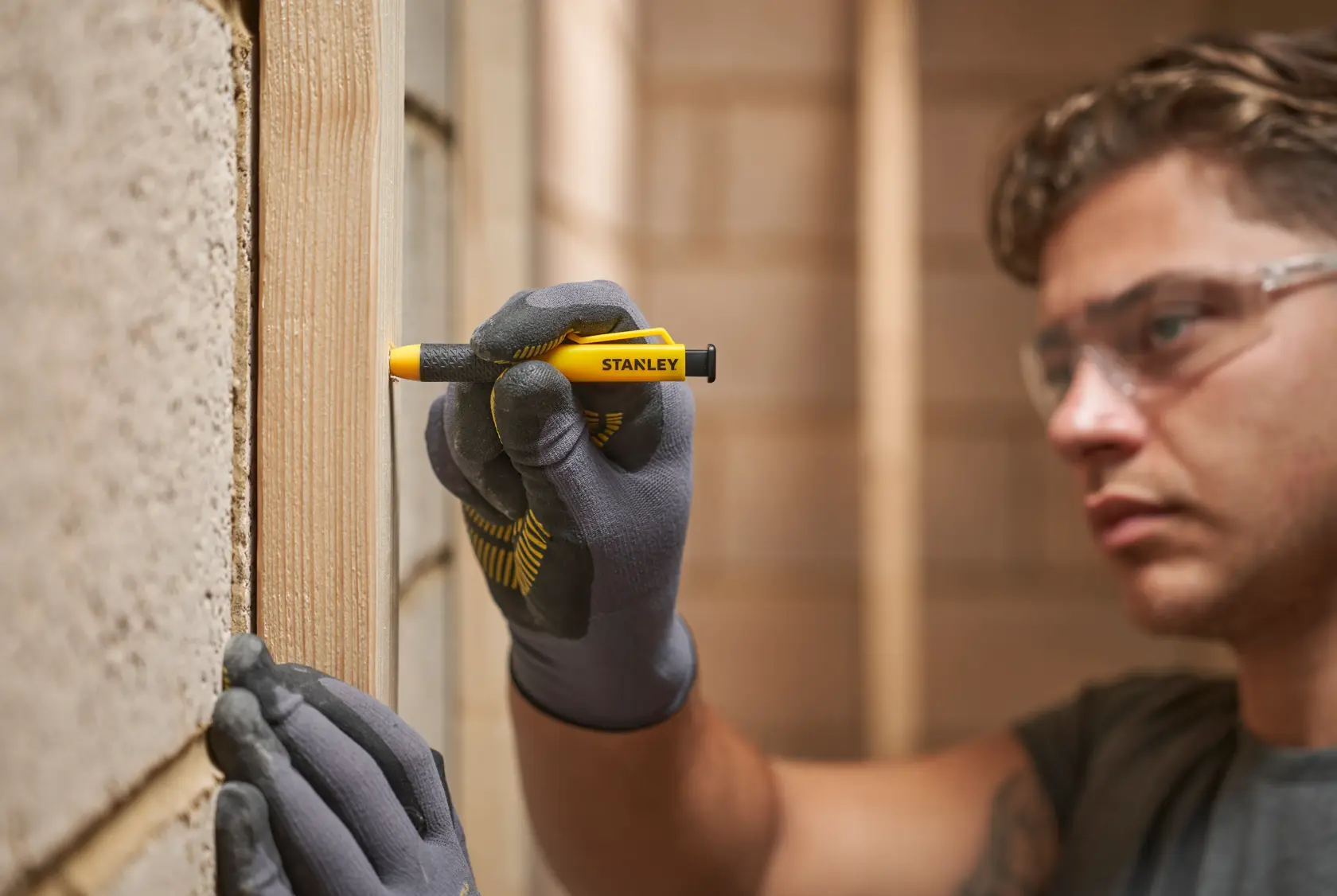 Person wearing gloves using a Stanley carpenter pencil to mark a wooden beam indoors.