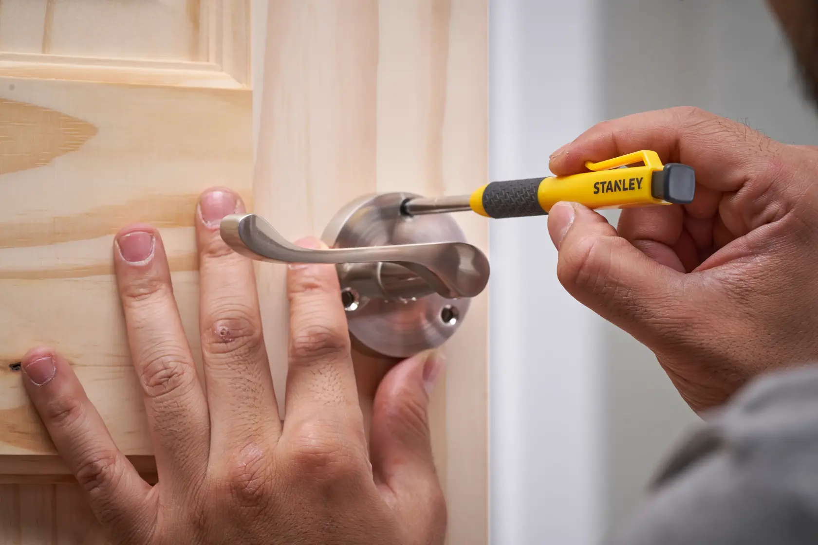 Person uses a Stanley screwdriver to tighten a screw on a metal door handle attached to a wooden door.