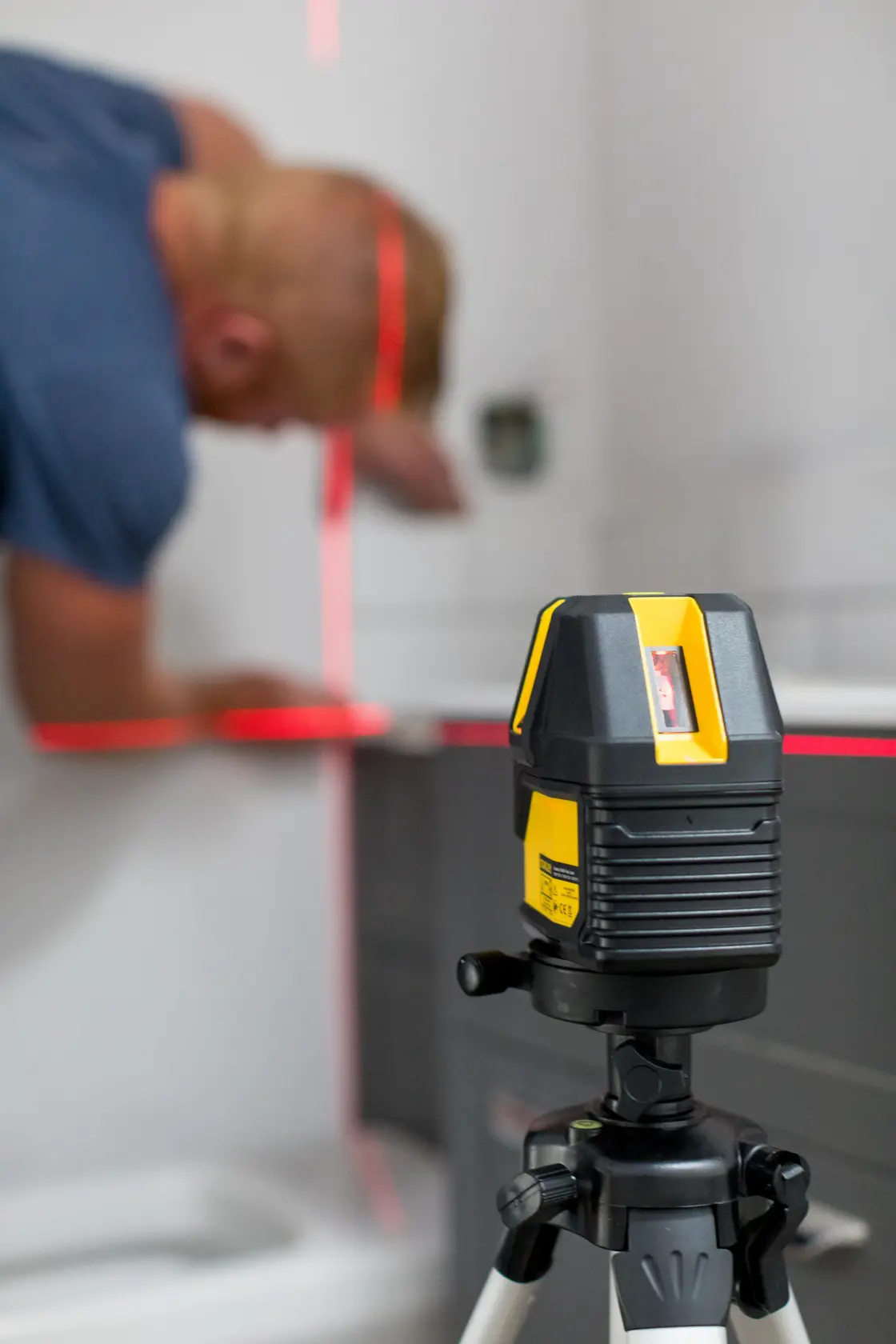 STANLEY STHT77514-1 cross line laser level on tripod projecting red lines, with person working in the background.