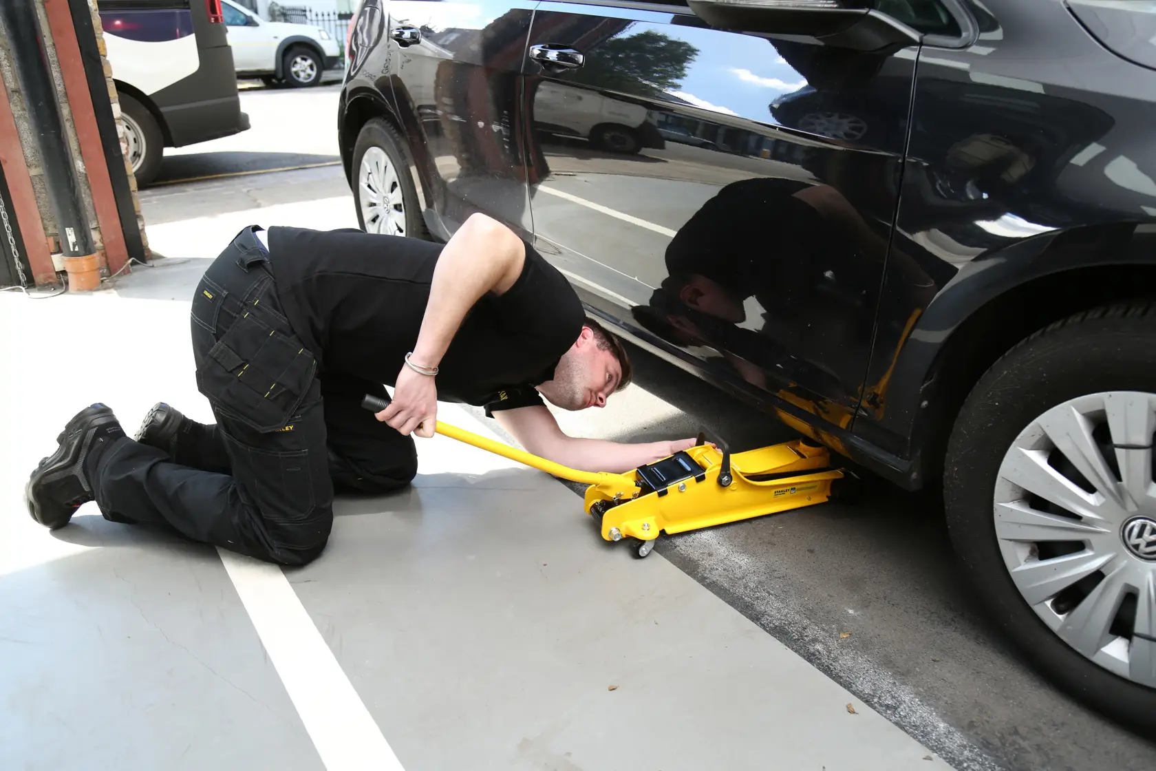 STANLEY STMT81251-1 yellow jack lifting a black car, technician kneeling beside it in workshop.