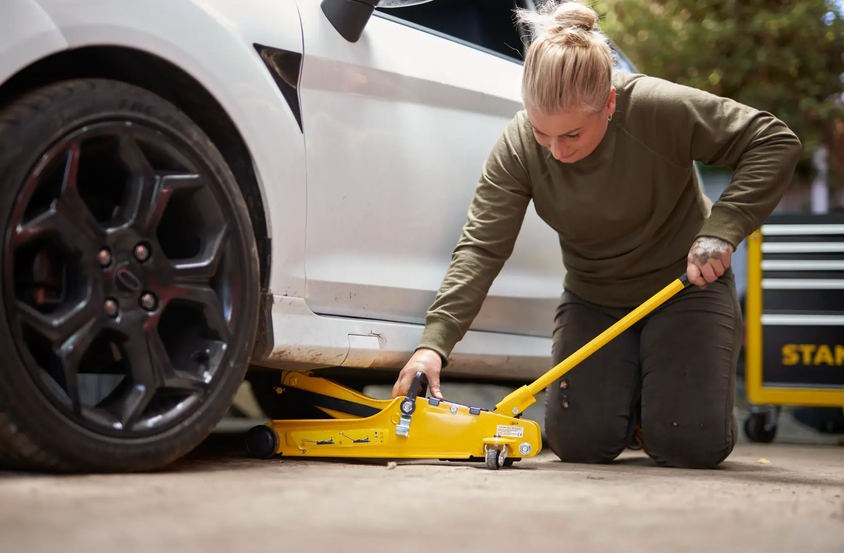 STANLEY STMT81251-1 yellow floor jack in use lifting a car, user kneeling beside, garage setting.