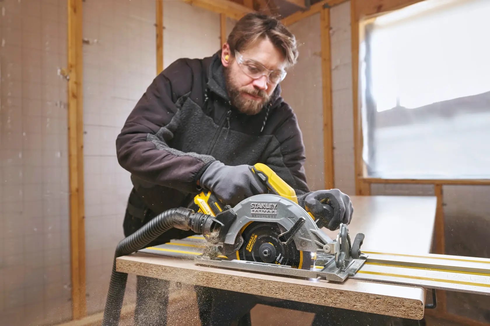 Photo of STANLEY STST140-XJ circular saw in use cutting wood, attached to a guide and dust hose, in a workshop.