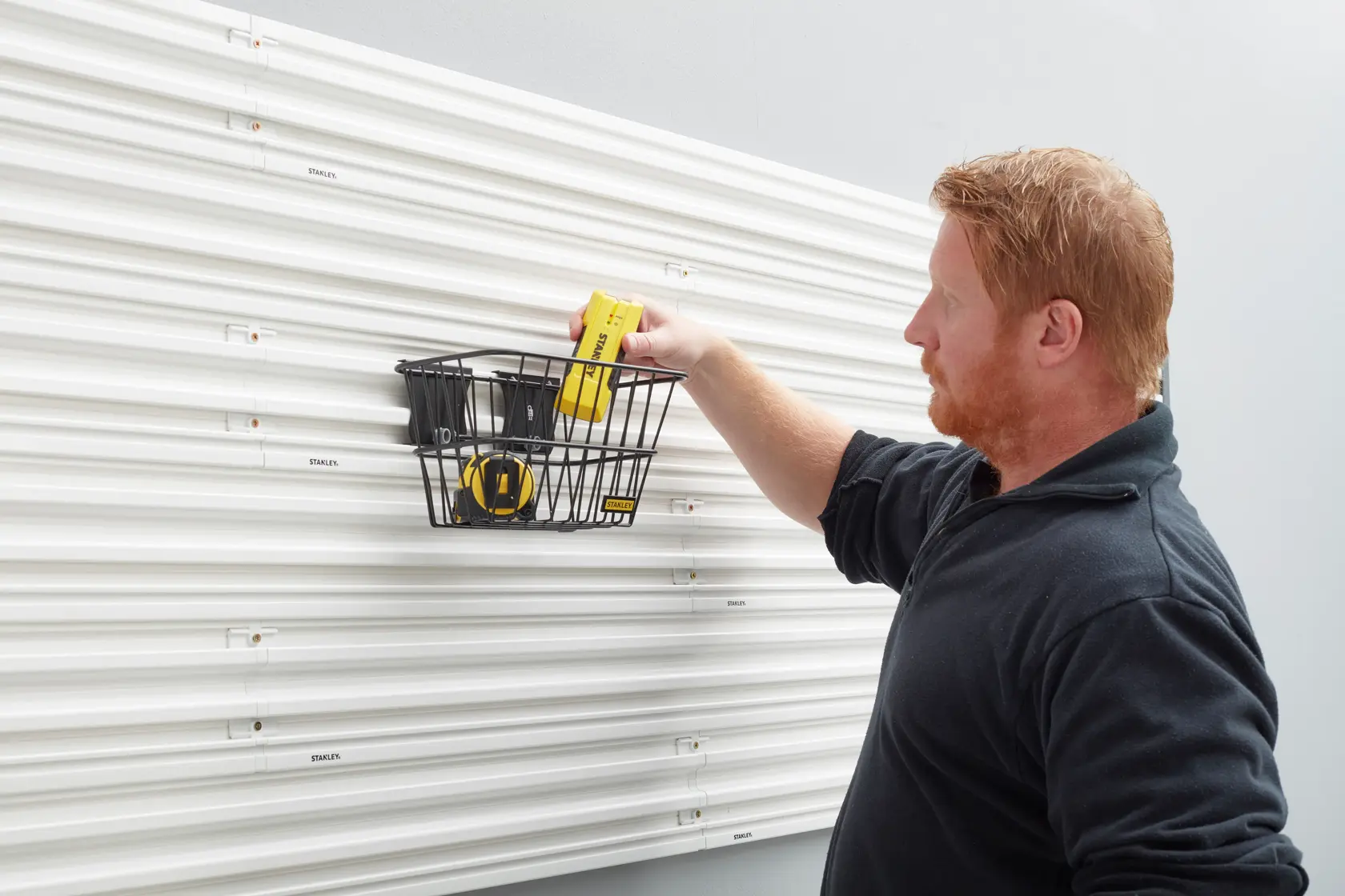 STANLEY STST82603-1 wall-mounted storage basket with tools on white slatwall, side angle photo.