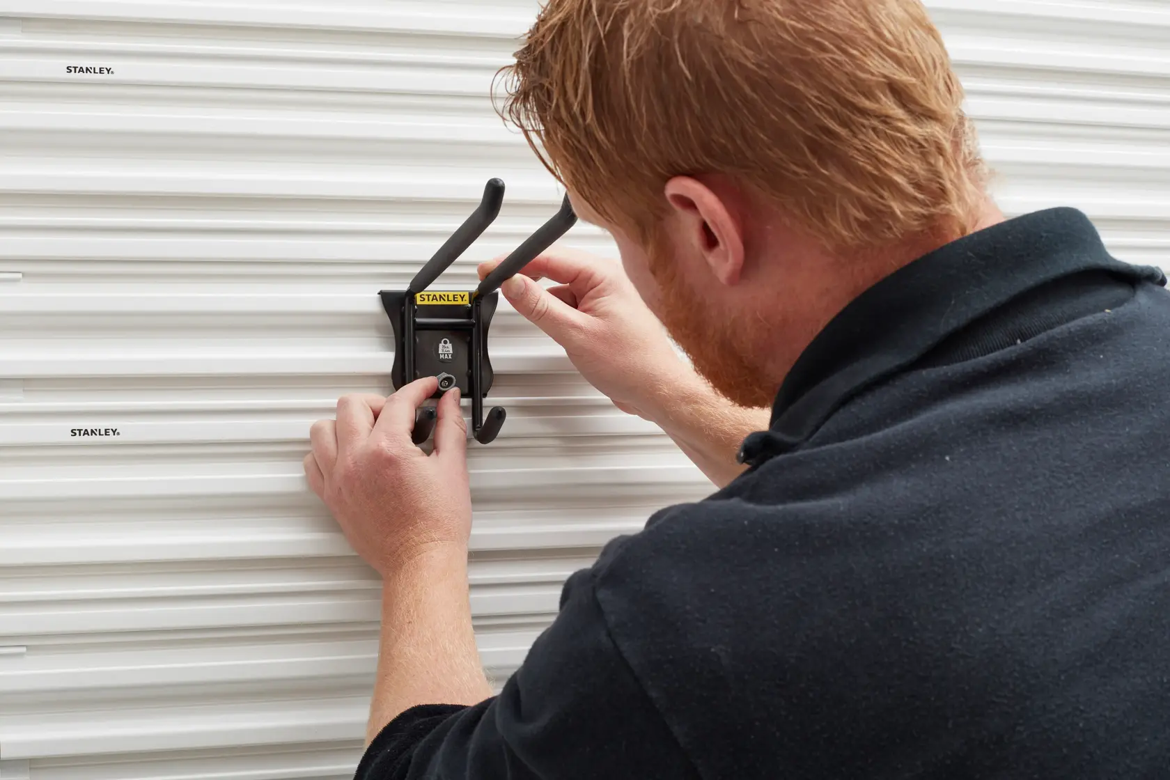 STANLEY STST82606-1 wall-mounted black storage hook being installed on ribbed white panel.