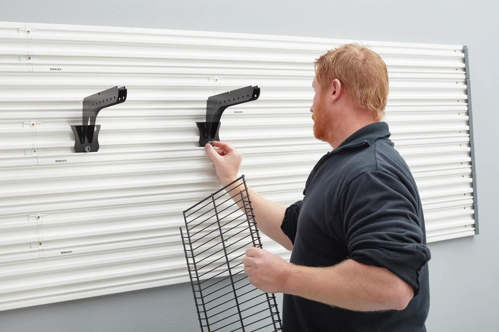 STANLEY STST82612-1 wall storage panel with black metal brackets, front angle, man holding wire shelf