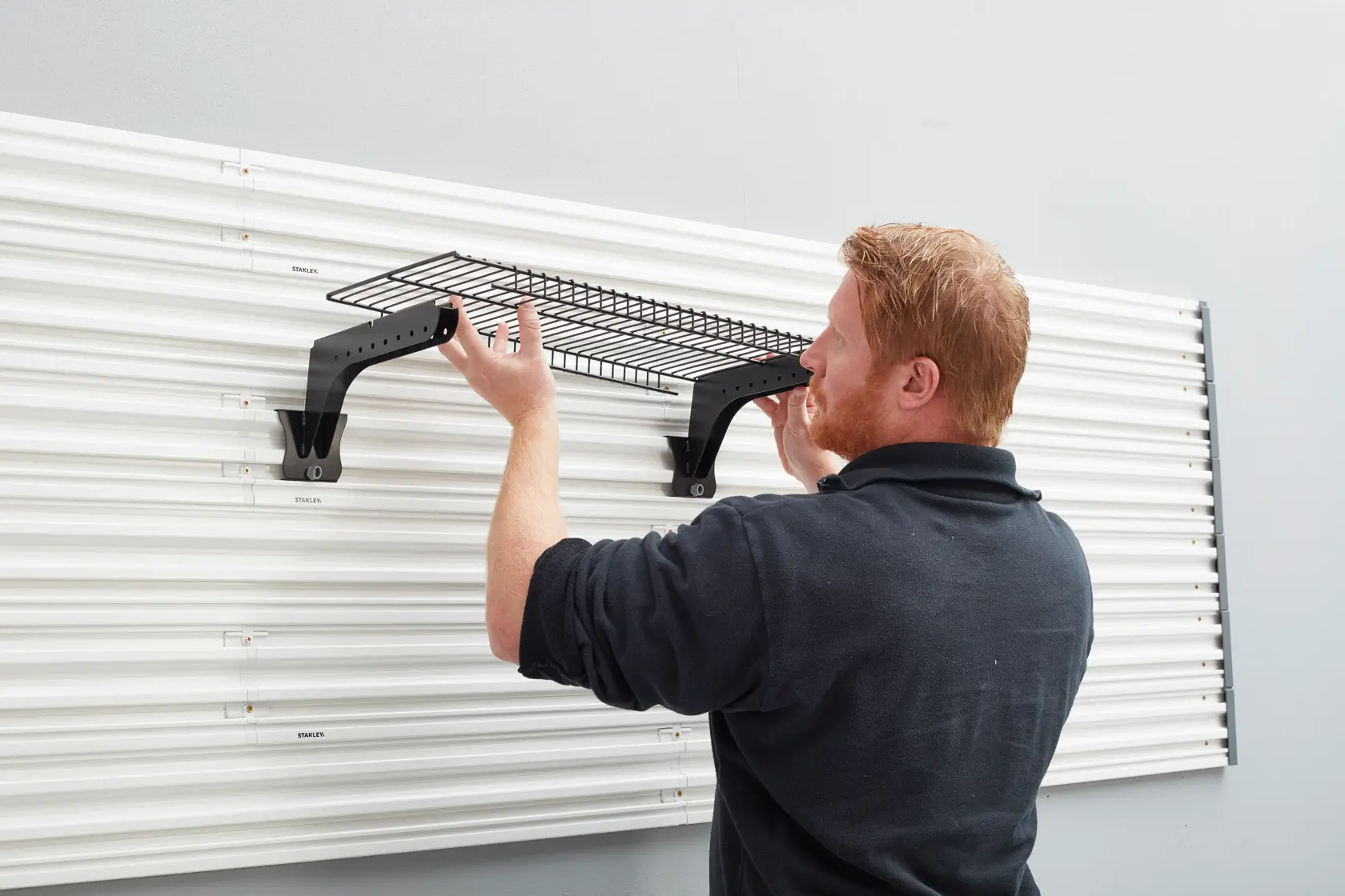Photo of a person installing a STANLEY STST82613-1 wall-mounted wire shelf on a white panel system.