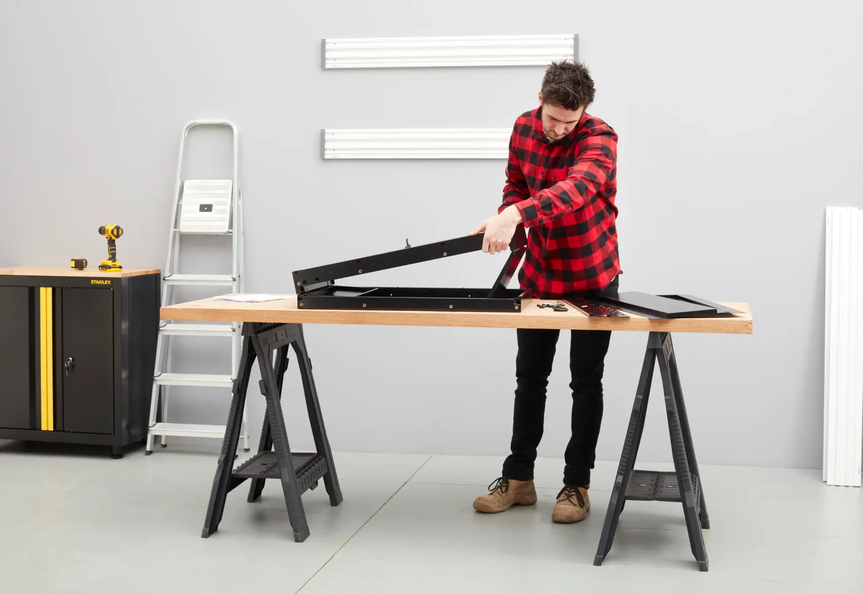 Photo of STANLEY STST97599-1 workbench in workshop, man assembling black metal frame on wood surface.